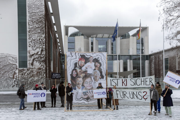 Erster Protest von Fairness Jetzt vor dem Kanzleramt. Das Bild zeigt einige Protestierende, die ein Plakat mit Menschen hervorheben, deren Augen durch das OpenAI Logo ersetzt wurde. Neben dran stehen Protestierende mit einem Plakat "Ist KI sicher für uns?"